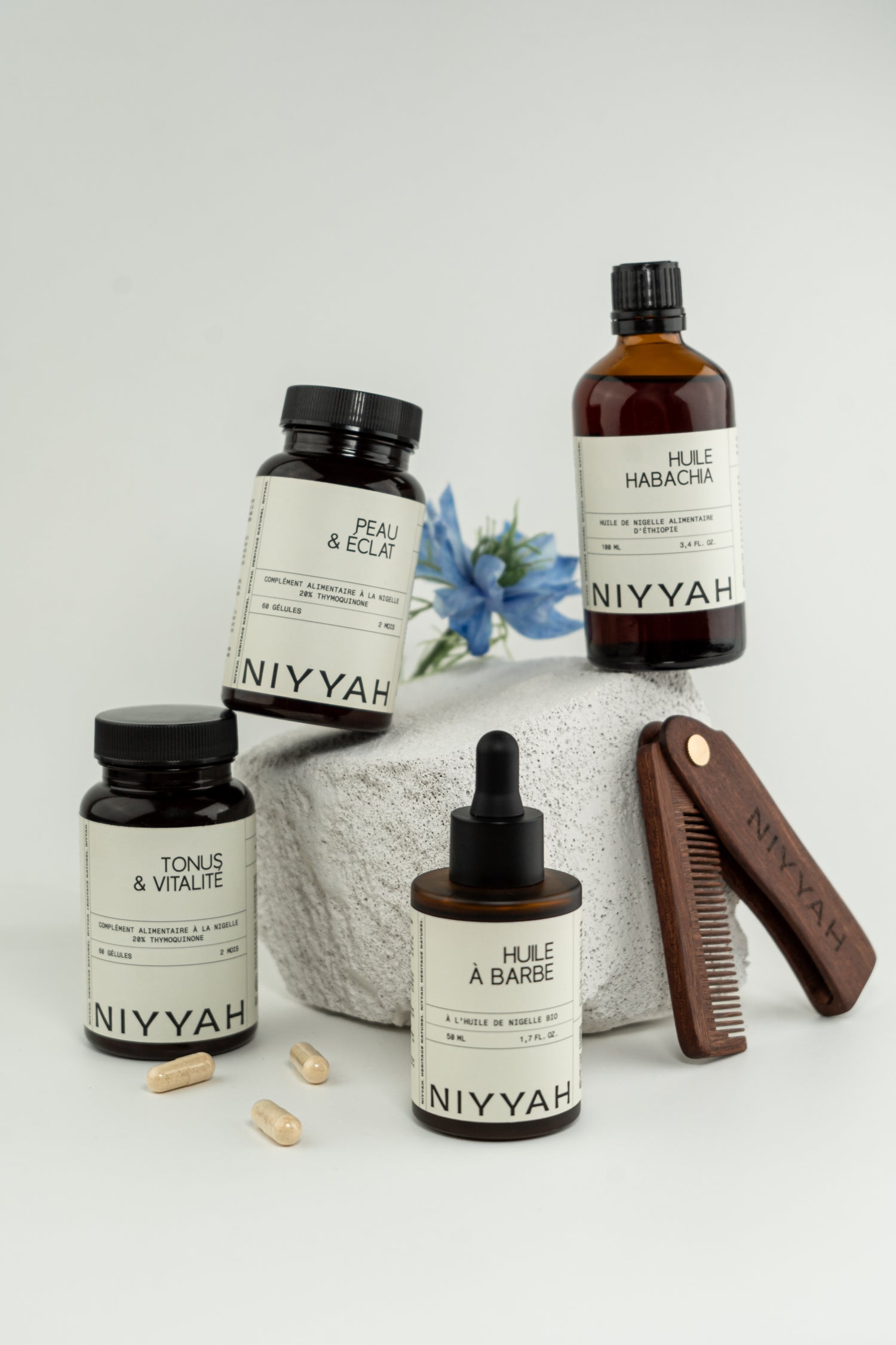 Produits NIYYAH à base de nigelle – huile de nigelle, huile à barbe, compléments alimentaires et peigne en bois, avec une fleur de nigelle en fond