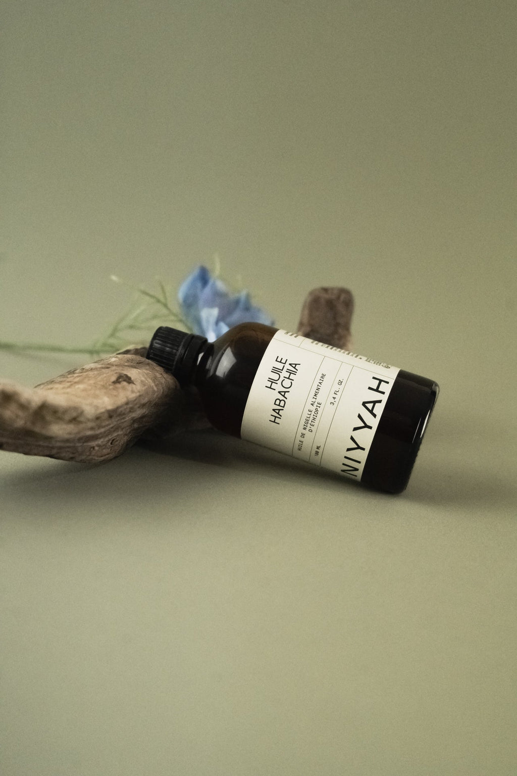 Flacon d'huile de nigelle Habacha alimentaire 100ml posé sur bois flotté avec fleur bleue nigella sativa en arrière-plan