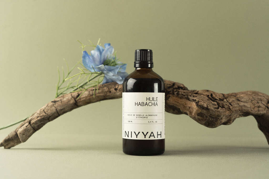 Huile de nigelle alimentaire Habacha 100ml d'Éthiopie posée sur branche naturelle avec fleur bleue de nigella sativa